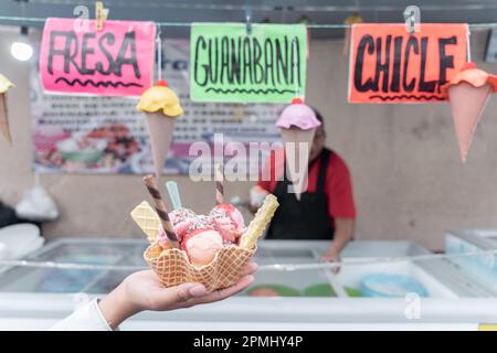 Eine Hand hält ein traditionell mexikanisch dekoriertes Eis vor einem Straßenmarkt mit Geschmacksschildern Stockfoto