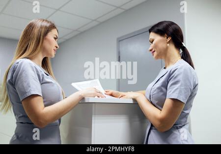 Schöne lächelnde weibliche Ärzte in medizinischer Uniform stehen in der Klinik in der Nähe des Fensters. Junge Krankenschwestern halten Papierbögen in ihren Händen. Sonniger Tag. Stockfoto