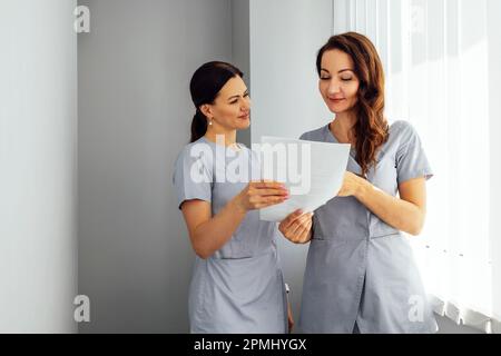 Schöne lächelnde weibliche Ärzte in medizinischer Uniform stehen in der Klinik in der Nähe des Fensters. Junge Krankenschwestern halten Papierbögen in ihren Händen. Sonniger Tag. Stockfoto