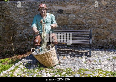 Eine bemalte Skulptur eines sitzenden Fischers mit einer Strickmütze, der Netz und Korb durch seine Hände gleiten lässt, steht an einer Steinmauer in der Altstadt von Kyrenia (türkisch: Girne), TRNK. Die Künstlerin ist Sevcan Çerkez, deren ausdrucksstarke Keramik- und Skulpturenwerke bereits in ihrem Atelier in Boğazköy/Agirda dokumentiert wurden. Stockfoto