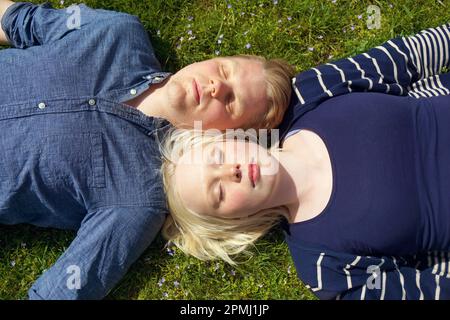 Junges Paar, das mit geschlossenen Augen auf Gras liegt und den Kopf Seite an Seite genießt und die Sonne genießt Stockfoto