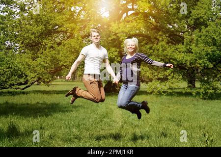 Junges Paar, das vor Freude hüpft, während es die Hände in der Natur hält - echte Menschen mit einem Leckfilter mit Sonneneinstrahlung Stockfoto