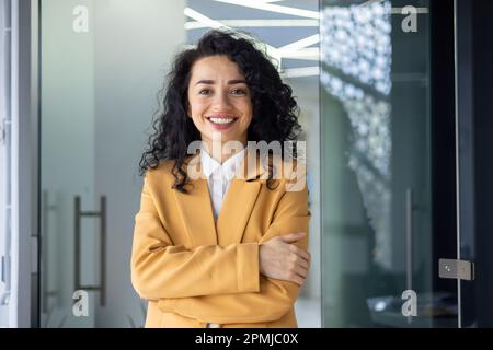 Porträt einer erfolgreichen Geschäftsfrau im Büro, lateinamerikanischer Boss im gelben Anzug, der lächelt und in die Kamera schaut, eine reife Mitarbeiterin mit lockigem Haar, die mit gekreuzten Armen steht. Stockfoto