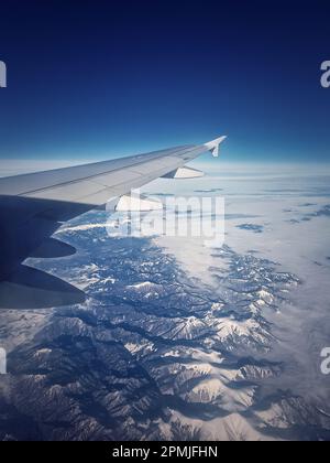 Flug über den schneebedeckten Gipfeln der Karpaten. Blaue Skyline und Flugzeugflügel, durch das Fenster gesehen, vertikaler Hintergrund Stockfoto