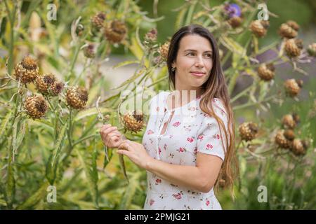 Junge, schöne Frau, die in der Nähe einer großen Klettenfabrik in einem alten, überwucherten Park steht Stockfoto