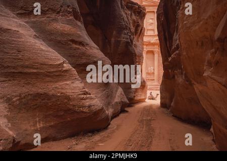 Al Siq Canyon in Petra, Jordanien, rosa Sandsteinwände auf beiden Seiten, unbekannte Person, die in Kamelentfernung sitzt, Schatztempel dahinter Stockfoto