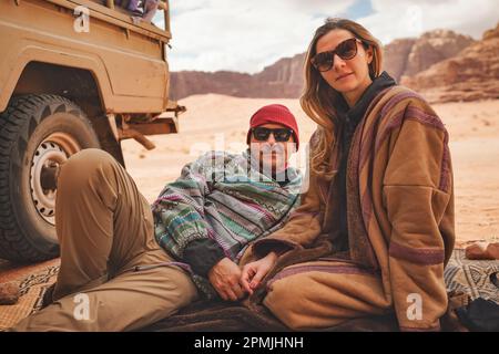 Mann und jüngere Frau tragen traditionellen Beduinen warmen Mantel - bisht - sitzen oder legen auf Boden Decke in der Nähe von Off-Road-Fahrzeug zurück, verschwommen Wadi Ru Stockfoto