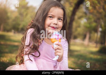 Süßes blondes, lockiges Mädchen, das an einem schönen Sommertag im Wald ein leckeres Eis isst, sie schaut in die Kamera, Kindertageskonzept, glückliches Mädchen Stockfoto