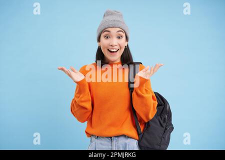 Glückliche asiatische Frau mit warmem Hut, klatscht von der Überraschung in die Hände, lacht und sieht erstaunt aus, aufgeregt von Smth, steht auf blauem Hintergrund Stockfoto