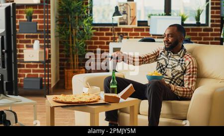 Lächelnde Person, die eine Flasche Alkohol in der Hand hält, Chips isst, Bier trinkt und Snacks im fernsehen isst. Ein junger Mann, der im Wohnzimmer Lieblingscomedy-Filme im Fernsehen sieht, Essen genießt. Stativaufnahme. Stockfoto