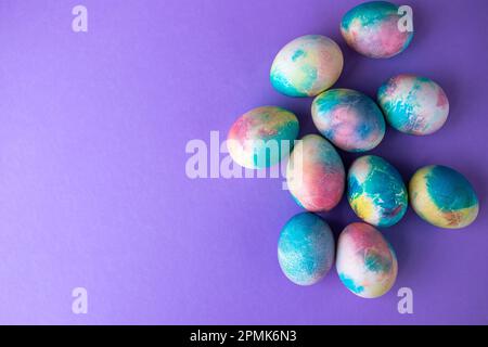 Ostergemisch mit bunten Eiern im Einkaufswagen, Holzhasen und Frühlingsblumen auf lila Hintergrund. Banner. Speicherplatz kopieren. Stockfoto