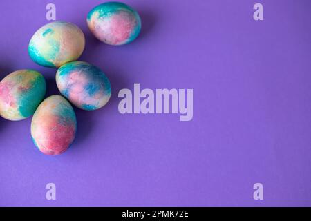 Ostergemisch mit bunten Eiern im Einkaufswagen, Holzhasen und Frühlingsblumen auf lila Hintergrund. Osterrahmen mit Eiern, die in lila C gestrichen sind Stockfoto