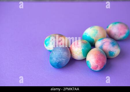 Ostergemisch mit bunten Eiern im Einkaufswagen, Holzhasen und Frühlingsblumen auf lila Hintergrund. Osterrahmen mit Eiern, die in lila C gestrichen sind Stockfoto