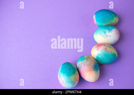 Ostergemisch mit bunten Eiern im Einkaufswagen, Holzhasen und Frühlingsblumen auf lila Hintergrund. Osterrahmen mit Eiern, die in lila C gestrichen sind Stockfoto