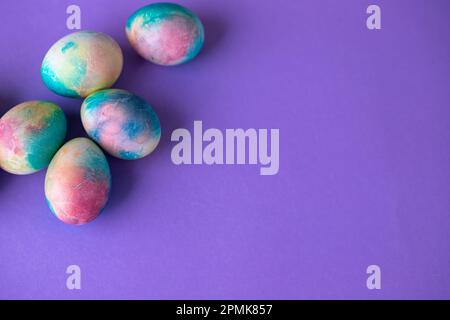 Ostergemisch mit bunten Eiern im Einkaufswagen, Holzhasen und Frühlingsblumen auf lila Hintergrund. Osterrahmen mit Eiern, die in lila C gestrichen sind Stockfoto