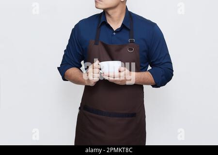 Asiatischer Mann trägt eine Schürze in lässiger, stylischer Kleidung, steht aufrecht und hält eine Tasse Kaffee oder Tee, eine ruhige und selbstbewusste Geste, kein Gesicht Stockfoto