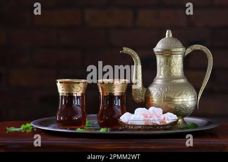 Eine klassische, feierliche, kunstvoll verzierte türkische Teekanne, zwei Gläser und traditionelle türkische Köstlichkeiten auf einem Tablett und einem Holztisch in sanfter stimmungsvoller Beleuchtung Stockfoto