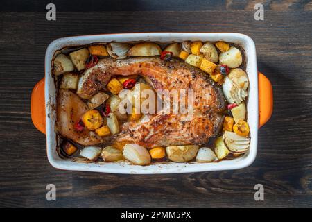 Gebackenes Lachsfilet mit Kartoffeln, Karotten, Zwiebeln, roten Paprika und Knoblauch in einer Keramikschüssel auf Holzhintergrund, Nahaufnahme Stockfoto