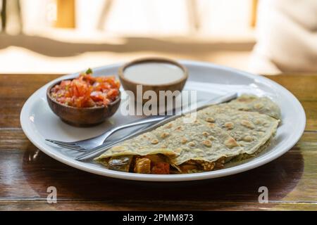 Quesadillas aus Spiruline Tortilla mit Cashewkäse, Auberginen, Tomaten und Sauerrahm und Tomatensalsa auf einem Holztisch im tropischen Café in Thailand, Stockfoto