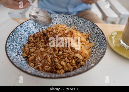 Schüssel mit Granulat und Kokosnuss- und Schokoladenchips für ein gesundes Frühstück. Männliche Hand mit einem Löffel und einem Teller mit Müsli in einem Café am Morgen, c Stockfoto