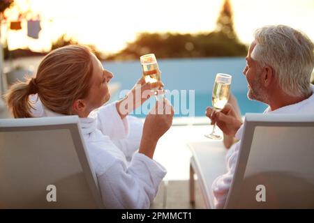 Auf einem romantischen Kurzurlaub. Rückblick auf ein reifes Paar, das Champagner trank, während es sich bei Sonnenuntergang auf den Liegestühlen entspannte. Stockfoto