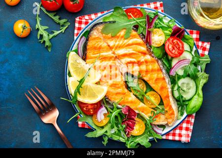 Gegrilltes Lachssteak mit Gemüsesalat mit Tomaten, Rucola und Salat auf dem Teller, blauer Tischhintergrund, Draufsicht Stockfoto