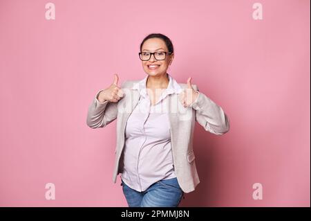 Fröhliche Schwangere mittleren Alters, die mit erhobenen Daumen lächelt und über einen isolierten pinken Hintergrund in die Kamera schaut Stockfoto