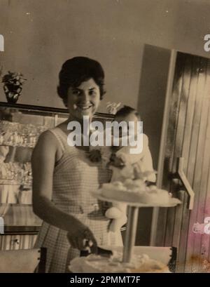 Rom, Italien Mai 1951: Das herzerwärmende Bild einer Mutter, die ihr Baby hält, während sie in einer nostalgischen 1950er-Kulisse einen Kuchen schneidet, um die geliebte Hungersnot zu feiern Stockfoto