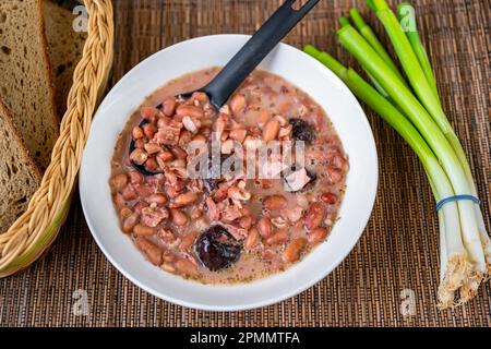 Leguminosen-Suppe aus Bohnen und Pflaumen auf weißem Teller, grüne Zwiebeln, Brot auf dem Tisch. Stockfoto