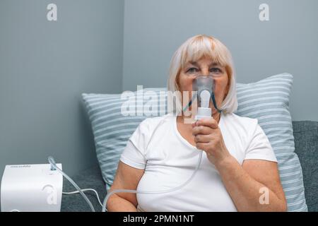 Porträt einer kranken Seniorin, die mit einem Inhalator sitzt. Eine ungesunde Frau, die zu Hause inhaliert, verwendet Vernebler und Inhalator für die Behandlung Stockfoto