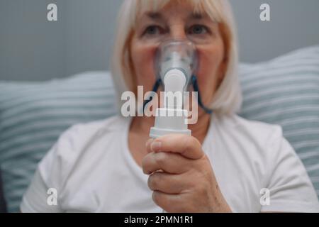 Porträt einer kranken Seniorin, die mit einem Inhalator sitzt. Eine ungesunde Frau, die zu Hause inhaliert, verwendet Vernebler und Inhalator für die Behandlung Stockfoto