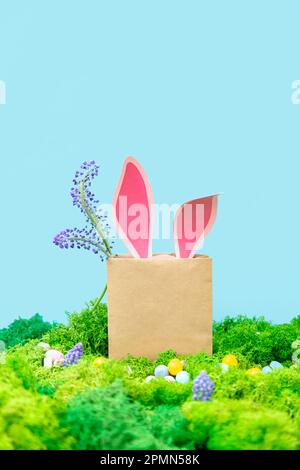 Die Ohren des rosa Kaninchens ragen aus dem Beutel. Frühling Grußkarte mit Gras und Blumen auf blauem Hintergrund. Ostersymbole. Stockfoto