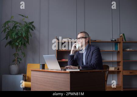 Erfolgreicher älterer, grauhaariger Geschäftsmann mit Brille, der auf einem Laptop arbeitet, mit einem Mobiltelefon an der Wand im Büro spricht Stockfoto