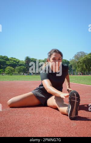 Vertikales Bild einer jungen latina, die sich das Bein und die Hüfte streckt und mit der Hand den Zeh berührt, sich auf der Laufstrecke aufwärmt, Vorderansicht, Kopierbereich. Stockfoto