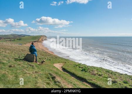 Eine Spaziergängerin und ihr Hund machen eine Pause entlang des South West Coast Pfades an der Jurassic Coast von Dorset mit Blick auf den Burton Freshwater Beach Stockfoto