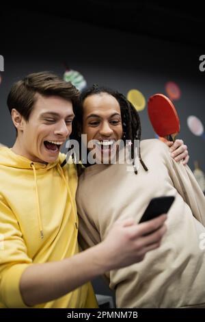 Positive zwischenrassische Freunde mit Tennisschläger, die im Spielclub ein Handy benutzen, Aktienbild Stockfoto