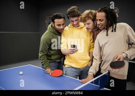 Lächelnde interrassistische Freunde, die ihr Smartphone in der Nähe von Tischtennis im Spielclub benutzen, Stock Image Stockfoto