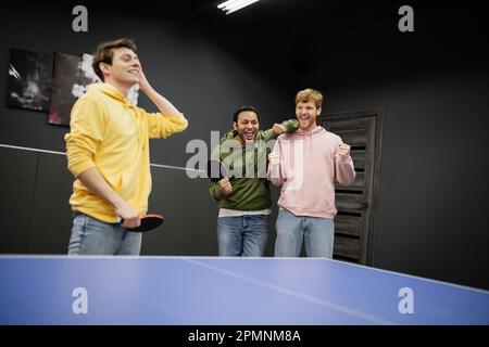 Aufgeregte, gemischtrassige Männer stehen neben einem verschwommenen Freund mit Tennisschläger im Spielclub, Aktienbild Stockfoto