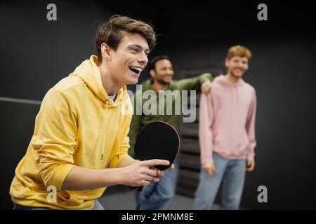 Fröhlicher Mann, der Tischtennis spielt, in der Nähe von verschwommenen Freunden im Spielclub, Aktienbild Stockfoto