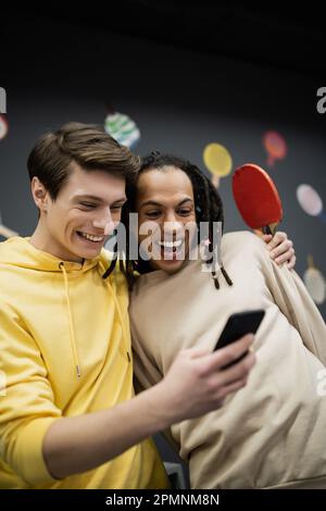 Ein lächelnder Mann, der ein Smartphone benutzt und einen Tennisschläger in der Nähe eines multirassischen Freundes im Spielclub hält, ein Aktienbild Stockfoto