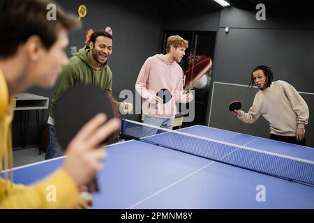 Aufgeregte Männer, die in der Nähe von Freunden stehen und Tischtennis spielen, im Spielclub, Aktienbild Stockfoto
