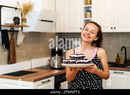 Brünette Frau steht in der Küche und hält Teller mit hausgemachten Cupcakes Stockfoto