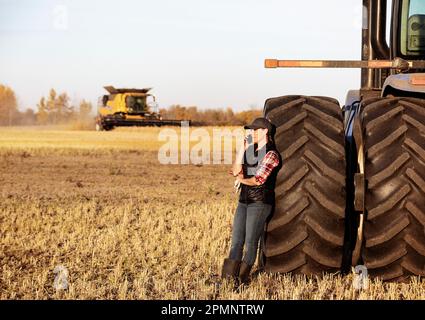 Eine reife Landwirtschaftsfrau, die sich an das Rad eines Traktors lehnt und mit ihrem Smartphone spricht, während ein Mähdrescher während der Herbsternte auf den Feldern arbeitet... Stockfoto