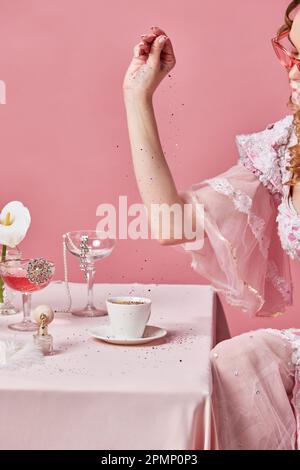 Abgeschnittenes Bild einer bezaubernden blonden Königin, die ein elegantes barockes Kleid trägt, an einem Tisch sitzt und Kaffee mit Glitzer über einem rosa Studio zubereitet Stockfoto