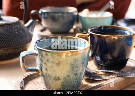 Tee zubereiten und servieren. Teekanne, grüner Tee, blauer Keramikbecher auf einem hölzernen Café-Tisch. Zwei handgemachte Töpferbecher am frühen Morgen. Trendiger Autor Stockfoto