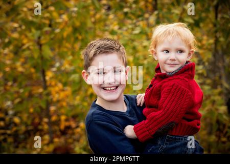 Porträt eines Jungen, der seinen kleinen Bruder im Herbst im Freien hielt; Edmonton, Alberta, Kanada Stockfoto