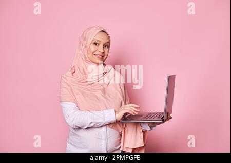 Wunderschöne, schwangere, arabische Moslem-Frau in pinkem Hijab, die online auf einem Laptop arbeitet, vor der Kamera lächelt, isolierter rosa Hintergrund Stockfoto