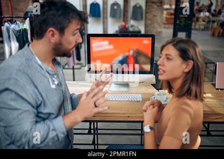 Junger Mann und junge Frau, die sich vor einem Desktop-Computer in einem Bekleidungsgeschäft unterhalten. Geschäftstreffen. Brainstorming. Manager und Mitarbeiter sprechen. Stockfoto