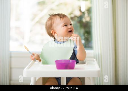 Süßes Baby isst erste feste Nahrung, Baby sitzt im Hochstuhl. Kind probiert Gemüse am Tisch und entdeckt neue Speisen. Gemütliche Kücheneinrichtung. Stockfoto