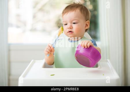 Süßes Baby isst erste feste Nahrung, Baby sitzt im Hochstuhl. Kind probiert Gemüse am Tisch und entdeckt neue Speisen. Gemütliche Kücheneinrichtung. Stockfoto